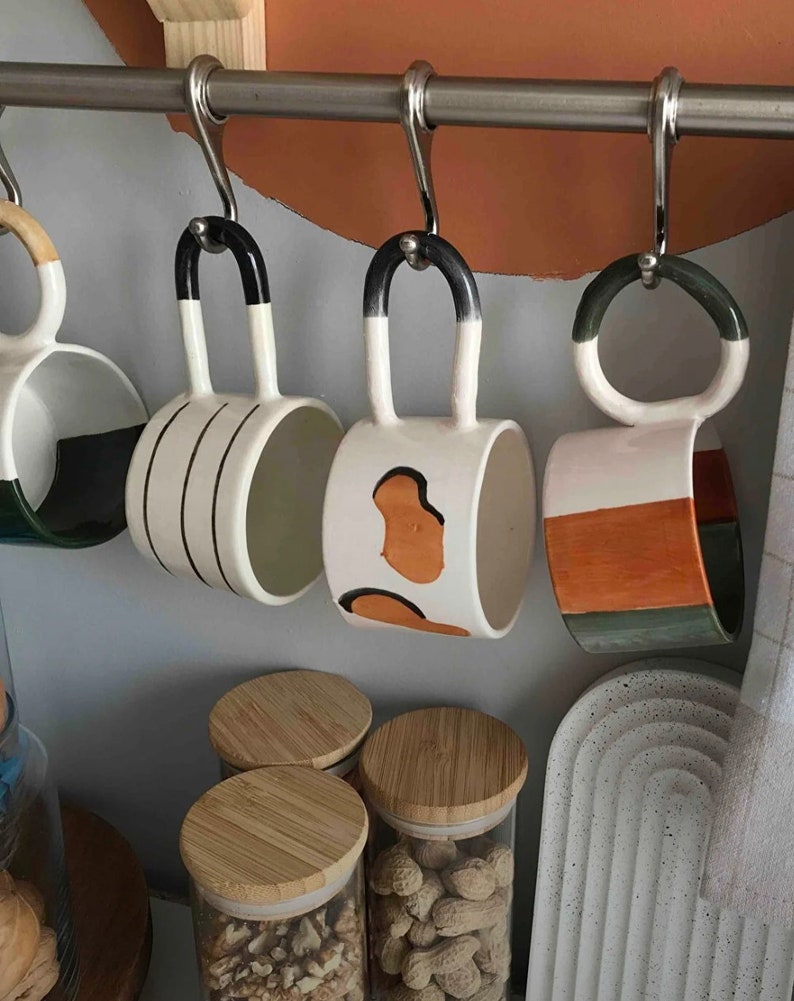 May include: Four ceramic mugs with different patterns hang on a metal rack. The mugs are white with black, orange, and green accents. The mugs are hanging on a metal rack with hooks.