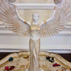 May include: White and gold winged statue of a woman holding two candle holders. The statue is standing on a round base and has a gold and white patterned dress.