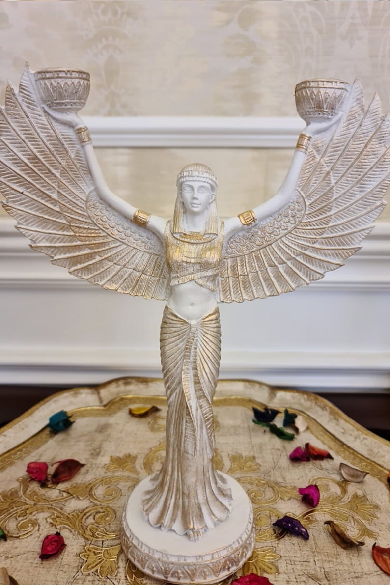 May include: White and gold winged statue of a woman holding two candle holders. The statue is standing on a round base and has a gold and white patterned dress.