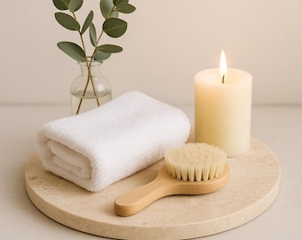 Könnte beinhalten: Eine Spa-Arrangement mit einem weißen, gerollten Handtuch, einer Bürste mit Holzgriff und einer brennenden Kerze. Eine kleine Glasvase mit Eukalyptuszweigen steht daneben auf einem runden, beigefarbenen Steintablett.