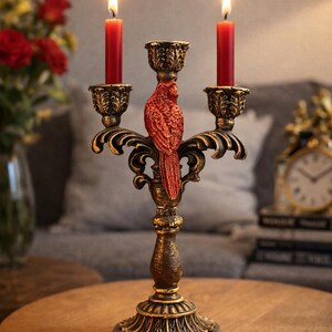 May include: Ornate, antique-style candelabra with three candle holders. The candelabra is bronze-colored with black accents and features a red bird sculpture. Two red candles are lit, casting a warm glow. The candelabra sits on a wooden table.