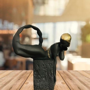 May include: Black and gold figurine of a scorpion with a curled tail and a human-like head. The figurine is sitting on a black square base.