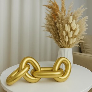 Könnte beinhalten: Eine goldene Kettengliedskulptur auf einem weißen Tisch. Die Skulptur besteht aus mehreren miteinander verbundenen Ringen, die jeweils eine glatte, abgerundete Oberfläche haben. Die Skulptur ist scharf, während der Hintergrund unscharf ist.