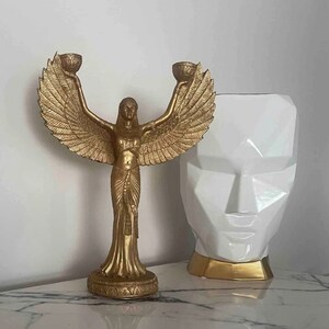 May include: Gold-toned decorative statue of a woman with wings holding two candle holders. The statue is on a white marble surface next to a white geometric vase with gold accents.