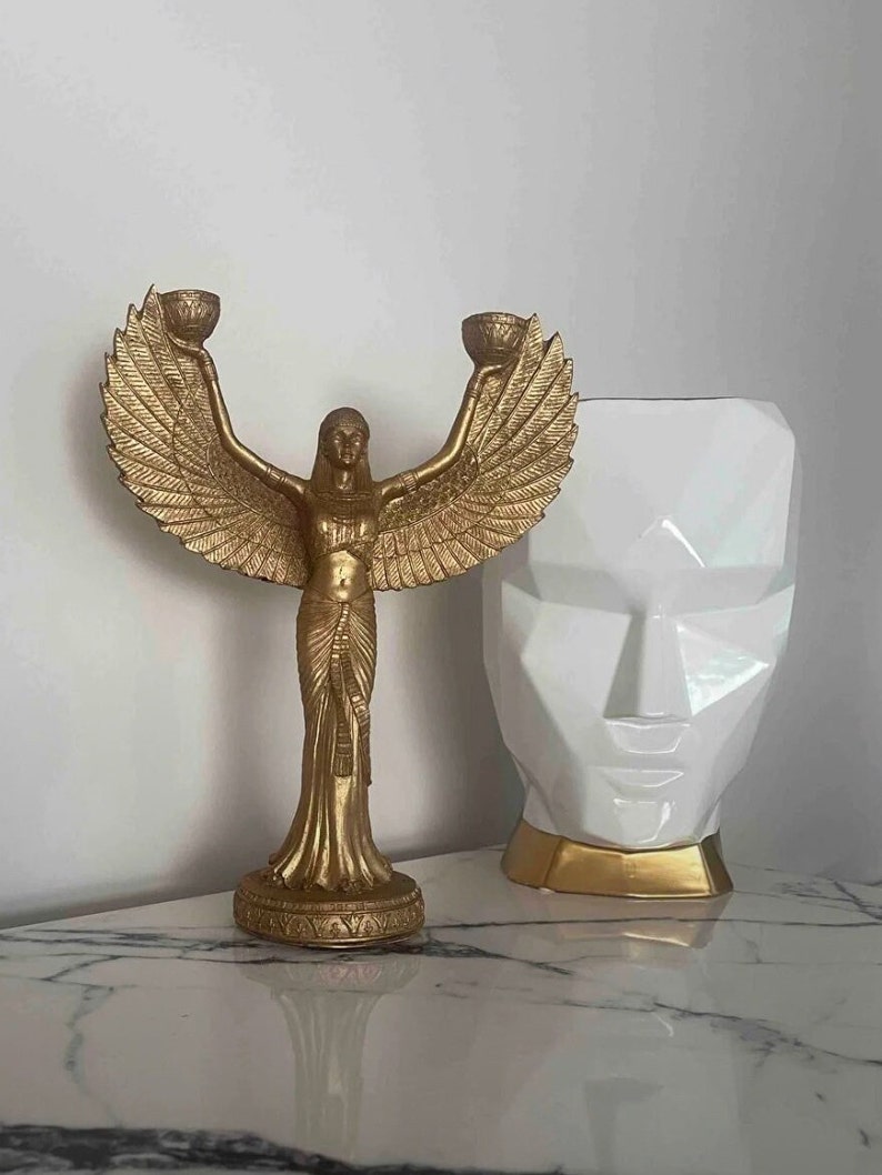 May include: Gold-toned decorative statue of a woman with wings holding two candle holders. The statue is on a white marble surface next to a white geometric vase with gold accents.