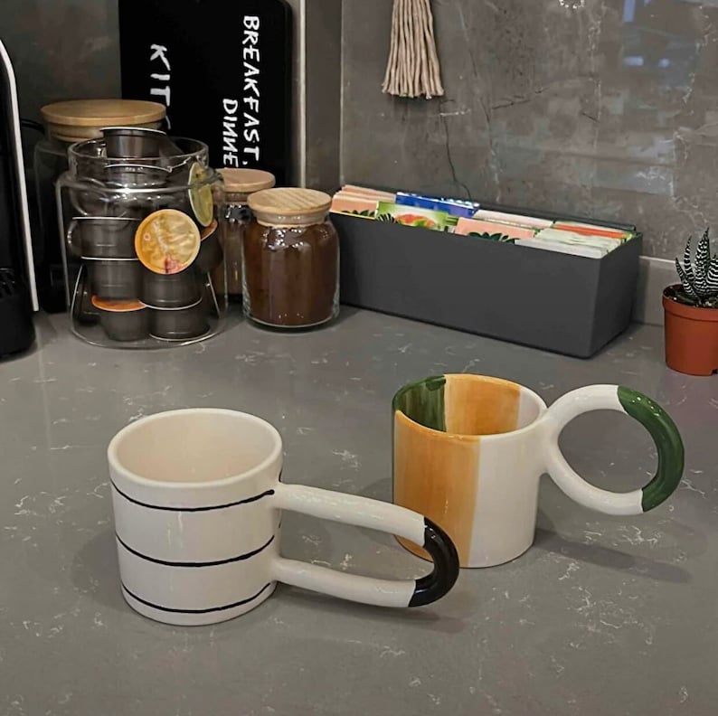May include: Two ceramic mugs with unique handles. One mug has a white base with black stripes and a black handle. The other mug has a white base with green, orange, and yellow stripes and a green handle.