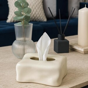 May include: A cream-coloured ceramic tissue box with a rectangular shape and a curved design. White tissues protrude from the top opening. The box is on a light-coloured table, with a vase and candle in the background.