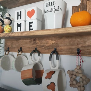 May include: A wooden shelf with two tiers. The top tier has a white mug with the text "Hello beautiful" and three wooden blocks that spell "HOME". The bottom tier has five hooks with four mugs hanging from them. The mugs are white with brown and green accents. There is a wooden star ornament hanging from the fifth hook.