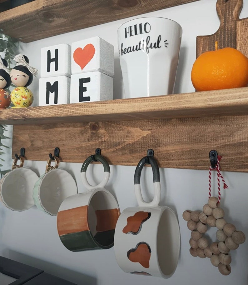 May include: A wooden shelf with two tiers. The top tier has a white mug with the text "Hello beautiful" and three wooden blocks that spell "HOME". The bottom tier has five hooks with four mugs hanging from them. The mugs are white with brown and green accents. There is a wooden star ornament hanging from the fifth hook.