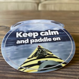 May include: Round sticker with a blue and white water background and a yellow kayak in the foreground. The text "Keep calm and paddle on" is printed in white on the sticker.