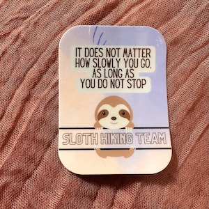 May include: A sticker with a cartoon sloth hanging from a branch. The sticker has a light blue background with a white border and the text "IT DOES NOT MATTER HOW SLOWLY YOU GO, AS LONG AS YOU DO NOT STOP SLOTH HIKING TEAM".