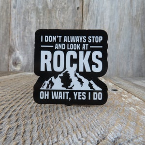 May include: Black sticker with white text that reads "I DON'T ALWAYS STOP AND LOOK AT ROCKS OH WAIT, YES I DO." The sticker has a mountain graphic and is on a wooden surface.