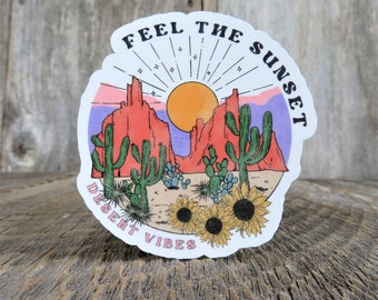 May include: A white round sticker with a desert sunset scene. The sticker features a red rock formation, cacti, and sunflowers. The text "Feel the Sunset" is written in a retro font at the top of the sticker. The text "Desert Vibes" is written in a retro font at the bottom of the sticker.