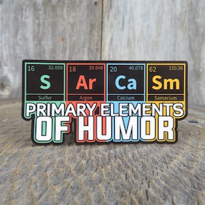 May include: A sticker with a black background and colorful squares featuring the chemical symbols for sulfur, argon, calcium, and samarium. The text "PRIMARY ELEMENTS OF HUMOR" is written in white letters below the squares.