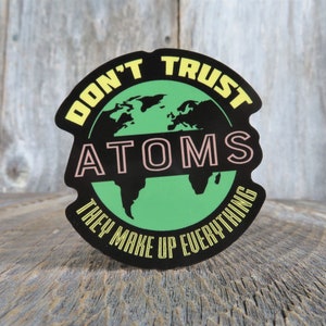 May include: A black and green round sticker with the text "Don't Trust Atoms They Make Up Everything". The sticker features a green outline of the Earth with the word "Atoms" in pink letters.
