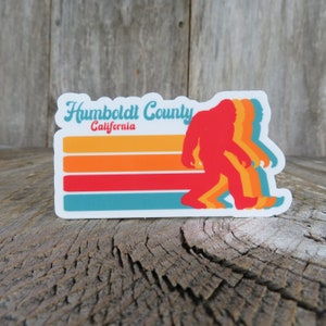 May include: A sticker featuring a red Bigfoot silhouette walking to the right, with layered orange, yellow, and blue outlines. The sticker has horizontal stripes in orange, red, and blue. Text reads "Humboldt County California" in a retro font.