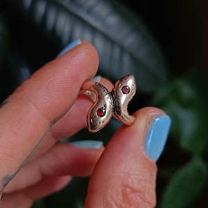 Könnte beinhalten: Ein goldfarbener Ring mit einem doppelten Schlangen-Design. Jeder Schlangenkopf hat ein kleines, rundes, rotes Edelsteinauge. Der Ring wird vor einem verschwommenen grünen Hintergrund gehalten, um die filigranen Details des Schmucks hervorzuheben.