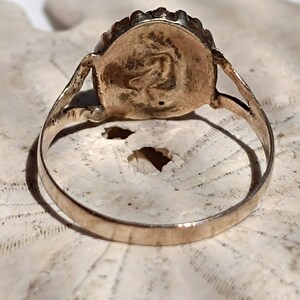 May include: A gold ring with a round, raised, textured design on the top. The ring is on a white, textured surface.