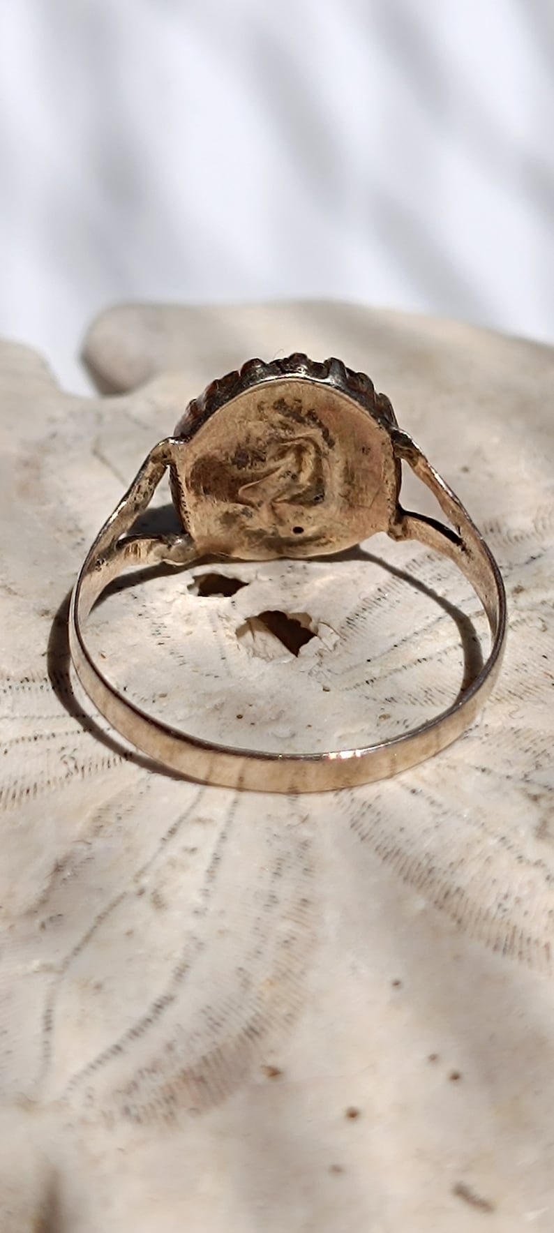 May include: A gold ring with a round, raised, textured design on the top. The ring is on a white, textured surface.