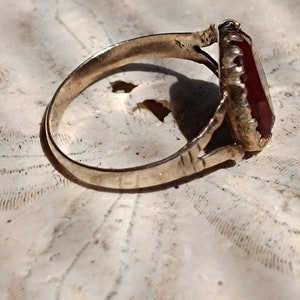 May include: A gold ring with a large, dark red gemstone set in a decorative, ornate setting. The ring is resting on a white, textured surface.