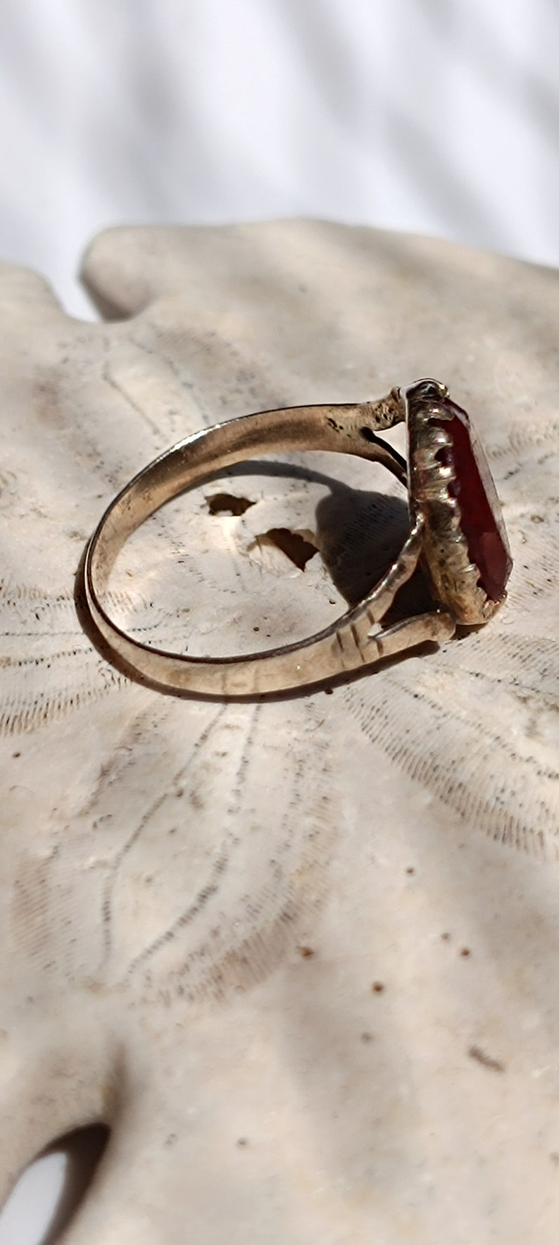 May include: A gold ring with a large, dark red gemstone set in a decorative, ornate setting. The ring is resting on a white, textured surface.