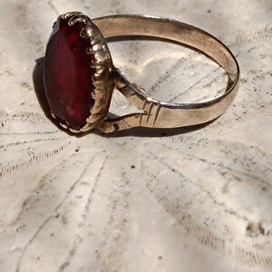 May include: A gold ring with a large, deep red gemstone set in a bezel setting. The ring is on a white background.