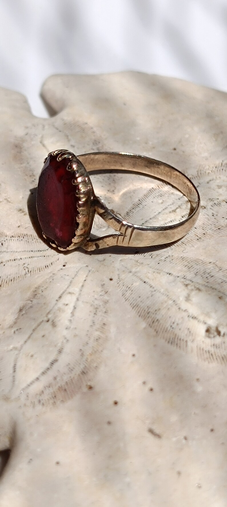 May include: A gold ring with a large, deep red gemstone set in a bezel setting. The ring is on a white background.