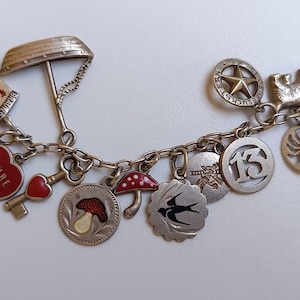 May include: A silver-toned charm bracelet featuring various dangling pendants, including a red heart with the text KEHRE WIEDER, a red mushroom, a swallow, a number 13, a star, a key, a boat, and several other circular and figurative metal charms.