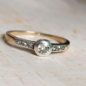 Può includere: Un anello in oro e argento con una grande gemma rotonda e trasparente al centro. La fascia presenta gemme più piccole su entrambi i lati della pietra centrale. L'anello è posto su una superficie chiara e strutturata.