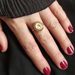 May include: A gold ring with a round, white shell cameo set in a gold bezel. The ring is on a finger with red nail polish.