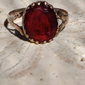 May include: A gold ring with a large, oval-shaped, dark red gemstone set in a gold bezel. The ring has a simple, delicate design.