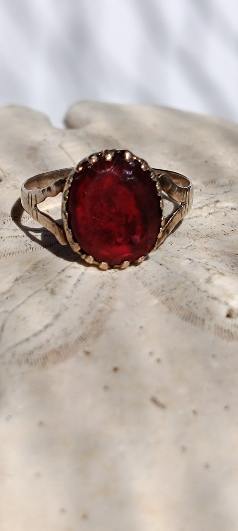 May include: A gold ring with a large, oval-shaped, dark red gemstone set in a gold bezel. The ring has a simple, delicate design.