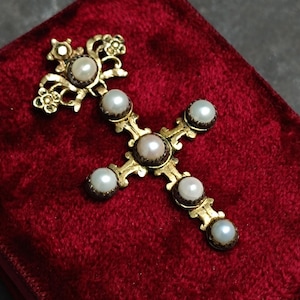 Pode incluir: Um pendente de cruz dourada com detalhes de pérolas, repousando sobre uma superfície de veludo vermelho. A cruz apresenta detalhes ornamentados e é adornada com várias pérolas redondas. O fundo é uma superfície escura e texturizada.