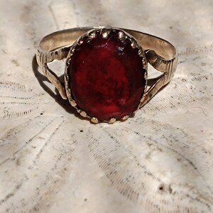 May include: A gold ring with a red oval gemstone set in a decorative bezel. The ring is on a white background.