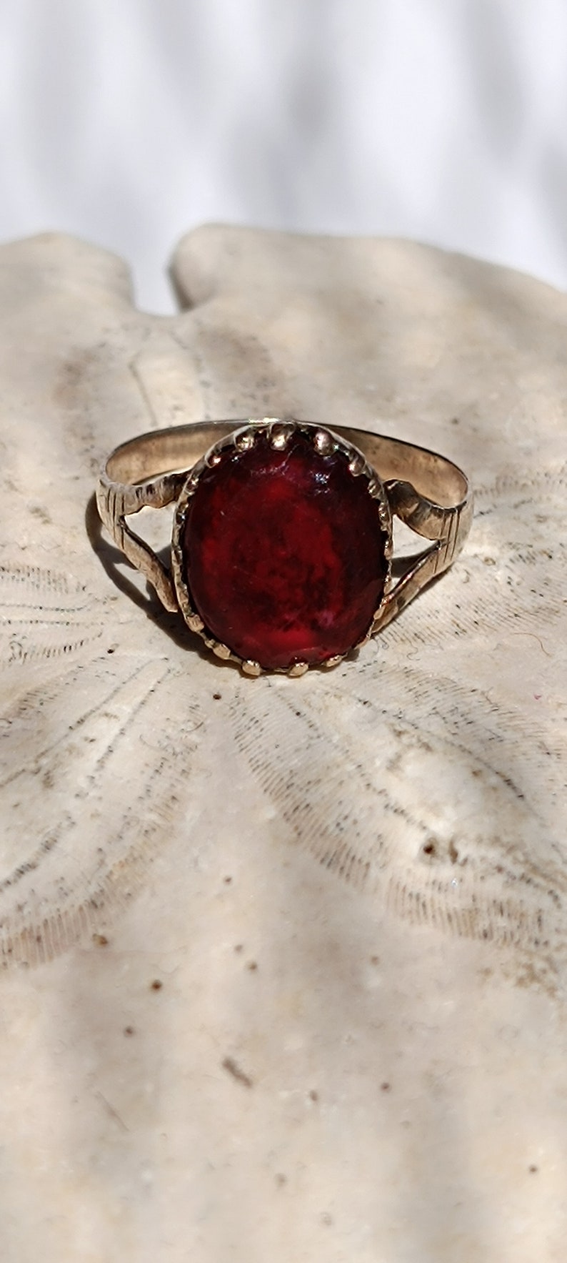 May include: A gold ring with a red oval gemstone set in a decorative bezel. The ring is on a white background.