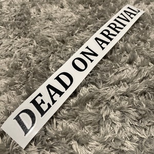 Op de afbeelding: Witte sticker met zwarte tekst die "DEAD ON ARRIVAL" luidt.