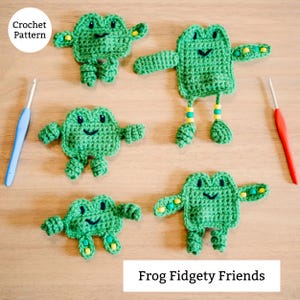 May include: Five green crochet frog toys with smiling faces and small arms and legs are displayed on a wooden surface. The frogs have yellow and green accents. The text "Frog Fidgety Friends" is at the bottom of the image. The text "Crochet Pattern" is in a circle at the top left.
