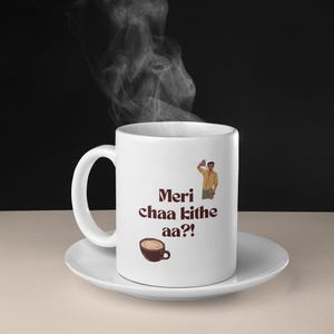 May include: White ceramic mug with a brown graphic that says "Meri chaa kithe aa?" and a cartoon illustration of a man holding a cup. The mug is sitting on a white saucer.