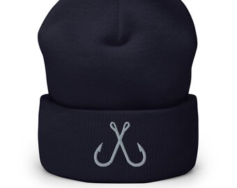 May include: A navy blue cuffed beanie featuring an embroidered light gray graphic of two crossed fishing hooks centered on the front fold. The knit hat has a structured crown and a thick, folded brim that displays the stitched design clearly.