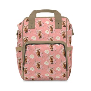 Könnte beinhalten: Ein rosa Rucksack mit einem Blumenmuster mit braunen Highland-Kühen. Der Rucksack hat ein großes Hauptfach und zwei Seitentaschen.
