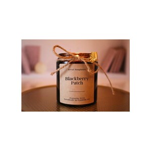 May include: A small, round glass jar candle with a brown label that reads "Blackberry Patch." The candle is tied with twine and has a gold-colored crown-shaped lid. The text "Koval Daughters" and "Handmade in Franklin, KY" are also visible.