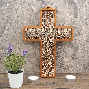 Könnte beinhalten: Ein Holzkreuz mit dem Vaterunser in Französisch darauf geschnitzt. Das Kreuz ist braun und der Text ist weiß. Das Kreuz hängt an einer grauen Wand.