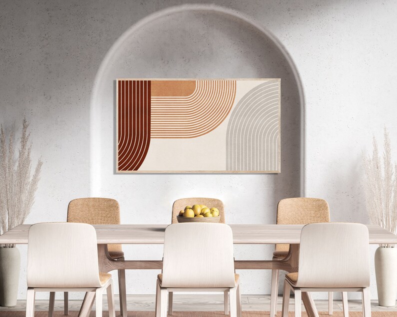 May include: A dining room with a white wall and a large archway. A framed abstract print with brown, orange, and gray geometric shapes hangs on the wall. A wooden table with six chairs is in the center of the room. A bowl of lemons sits on the table.