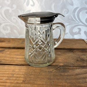 May include: A clear glass creamer pitcher with a diamond pattern and a metal lid. The pitcher has a handle and a spout. The lid is black and silver. The creamer pitcher is sitting on a wooden surface.
