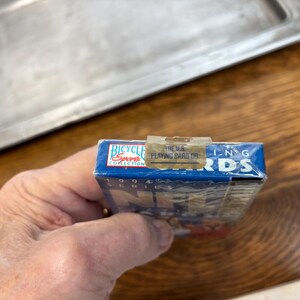 Pode incluir: Um baralho de cartas Bicycle Sports Collection azul da s&eacute;rie de 1994. O baralho &eacute; selado em pl&aacute;stico e segurado por uma m&atilde;o. A parte superior da caixa tem o texto "THE U.S. PLAYING CARD CO."