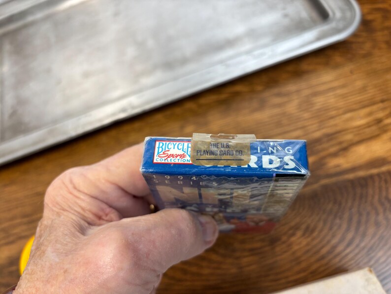 Pode incluir: Um baralho de cartas Bicycle Sports Collection azul da s&eacute;rie de 1994. O baralho &eacute; selado em pl&aacute;stico e segurado por uma m&atilde;o. A parte superior da caixa tem o texto "THE U.S. PLAYING CARD CO."