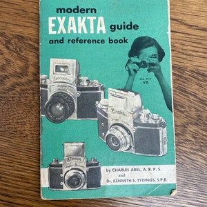 May include: A green book cover with the title "modern EXAKTA guide and reference book". The cover features a woman holding a camera and three different cameras. The text "the new VX" is printed below the woman.