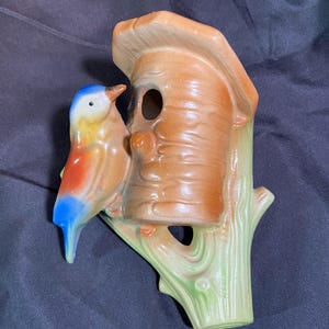 May include: A ceramic birdhouse planter with a blue, yellow, and red bird perched on a branch. The birdhouse is brown with a green branch.