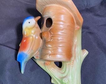 May include: A ceramic birdhouse planter with a blue, yellow and red bird perched on a branch. The birdhouse is brown with a green branch.