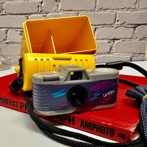 May include: A vintage Le Mini film camera with a blue and gray body, black lens, and colorful accents. The camera sits on a red book with the text "AMPHOTO". A yellow case is behind the camera.